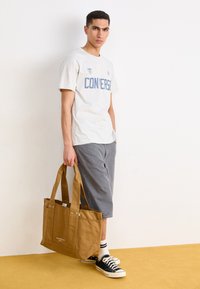 Gray cotton t-shirt with blue "CONVERSE" print, gray shorts, brown canvas tote bag, black sneakers, and white striped socks on a yellow floor.