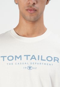 Nahaufnahme einer Person, die ein weißes T-Shirt trägt, auf dem in blauer Schrift "TOM TAILOR THE CASUAL DEPARTMENT 1962" auf der Brust gedruckt ist.