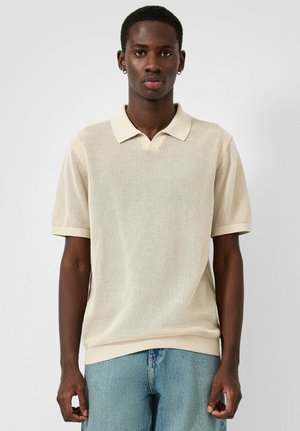 Junger Mann mit kurzen Haaren und Ohrringen, der ein beiges, kurzärmeliges Strick-Poloshirt und hellblaue Jeans trägt, steht vor einem weißen Hintergrund.