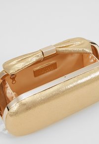 Guldfärgad metallclutch med rosettbetoning, hårt skal och slät yta; har en mjuk, ljus beige insida och magnetisk stängning.