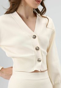 Roomkleurige cropped cardigan met een V-hals, voorzien van drie metalen knopen en een geribbelde structuur. Lange mouwen en een aansluitend design.