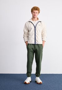 Chaqueta de forro polar crema con un patrón geométrico, cremallera azul marino, pantalones de chándal verdes y zapatillas blancas con suelas negras y marrones.