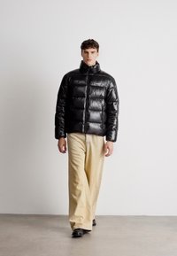 Μαύρο πουπουλένιο μπουφάν με ψηλό γιακά και κλείσιμο με φερμουάρ, συνδυασμένο με ανοιχτό μπεζ παντελόνι με φαρδιά γραμμή. Λείο, γυαλιστερό υλικό.
