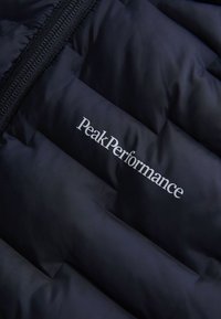 Tamno plava puffer jakna s prošivenim sekcijama, s sjajnom površinom. Logo "PeakPerformance" u bijeloj boji na donjem dijelu.