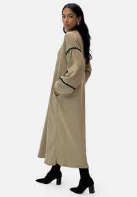 Khaki kordflorklänning med långa ärmar, prydd med svarta detaljer och sidfickor, bärs med svarta klackstövletter.