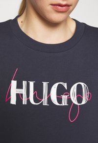 Sudadera azul marino con cuello de canalé, que presenta un audaz texto en blanco "HUGO" y un acento en espiral rosa. Textura de tela suave visible.