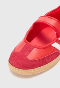 Red sports shoe featuring a combination of smooth synthetic material and soft suede, with a curved design and contrasting white stripes.