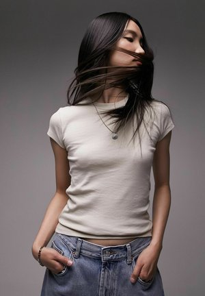 Jeune femme aux longs cheveux noirs tournant sur elle-même, vêtue d'un T-shirt blanc ajusté, d'un jean bleu, d'un collier et d'un bracelet, sur un fond gris uni.