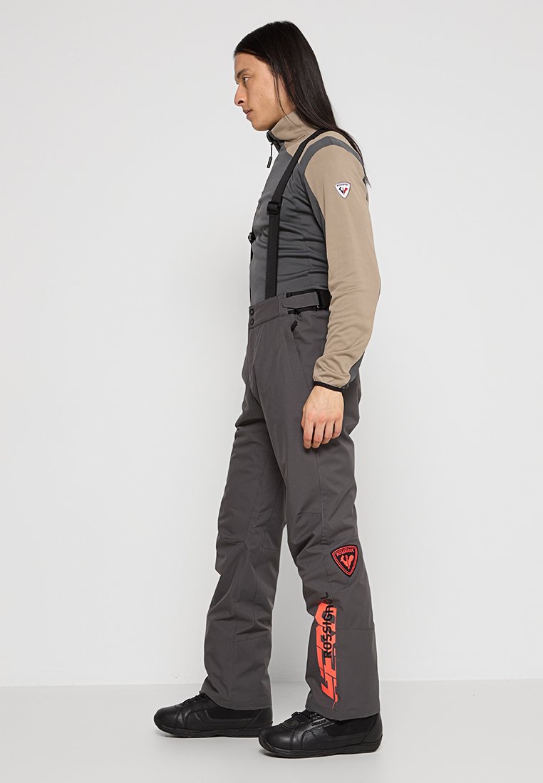 Szare ogrodniczki narciarskie z regulowanymi czarnymi ramiączkami, z czerwonymi logo i akcentami. Wykonane z trwałego materiału, zaprojektowane do aktywności zimowych.