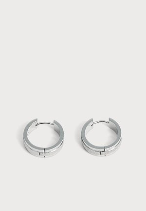 Paire de petites boucles d'oreilles en argent en forme de créoles avec un motif subtilement texturé, placées sur un fond clair uni.