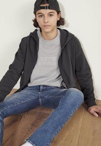 Adolescent assis sur un sol en bois contre un mur blanc, portant une casquette noire, un sweat à capuche noir, un t-shirt gris "Édition Limitée" et un jean bleu.