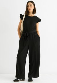 Black textured jumpsuit with short ruffle sleeves, tied waistband, and wide-leg fit. Paired with black sneakers and a cream shoulder bag.