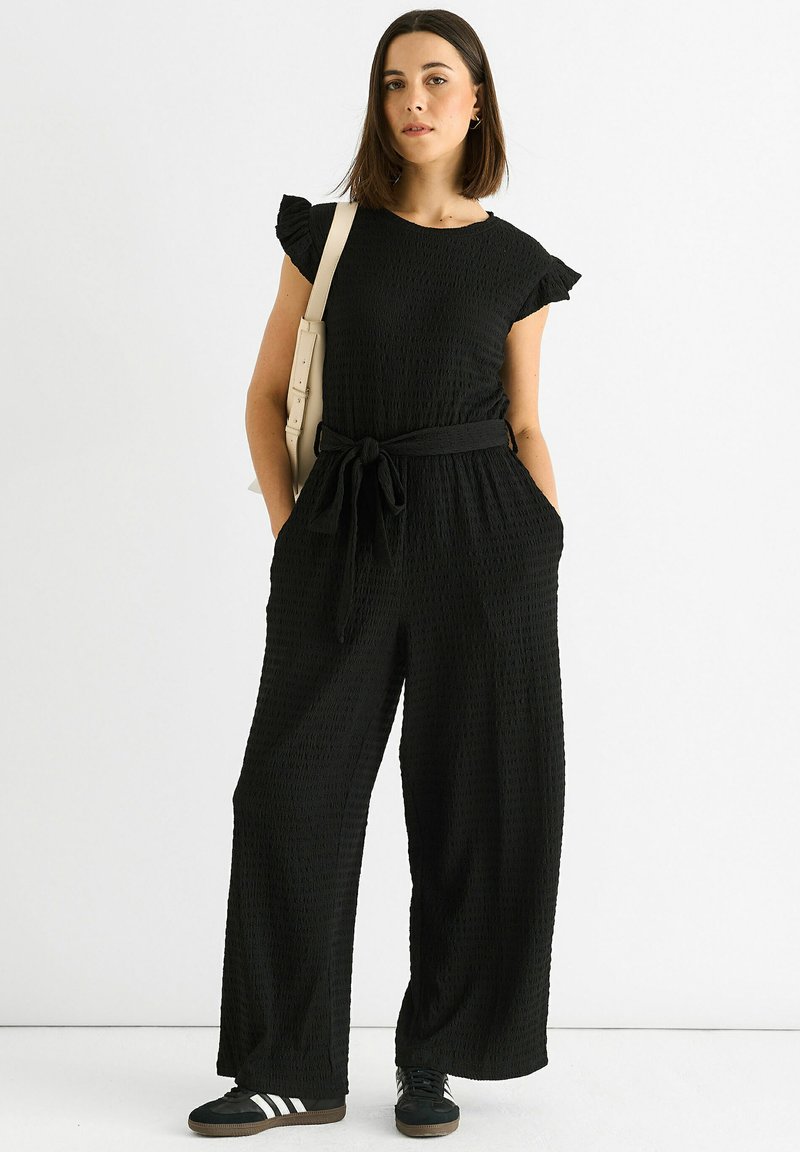 Black textured jumpsuit with short ruffle sleeves, tied waistband, and wide-leg fit. Paired with black sneakers and a cream shoulder bag.
