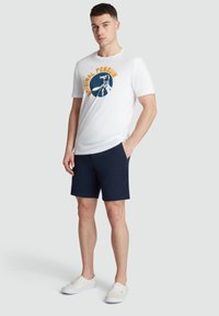 Weißes Baumwoll-T-Shirt mit blauer, runder Grafik und "Original Penguin"-Text, kombiniert mit marineblauen Shorts und weißen Turnschuhen.