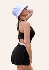 Top de baño negro con corte en la espalda y lazo ajustable. Falda negra con cinturilla blanca. Sombrero tipo cubo blanco. Tejido suave, diseño deportivo.