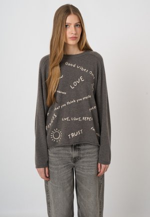 Woman with long light brown hair wears gray sweater with positive words like Love, Trust, and Good Vibes Only, paired with faded gray jeans.