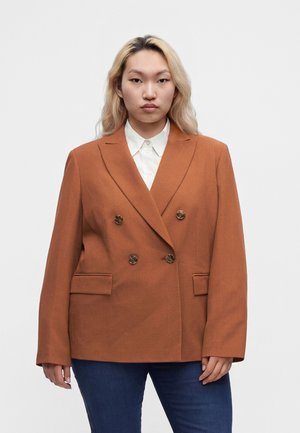 Personne aux longs cheveux blonds porte un blazer croisé couleur rouille sur une chemise blanche et un jean bleu, debout devant un fond uni.
