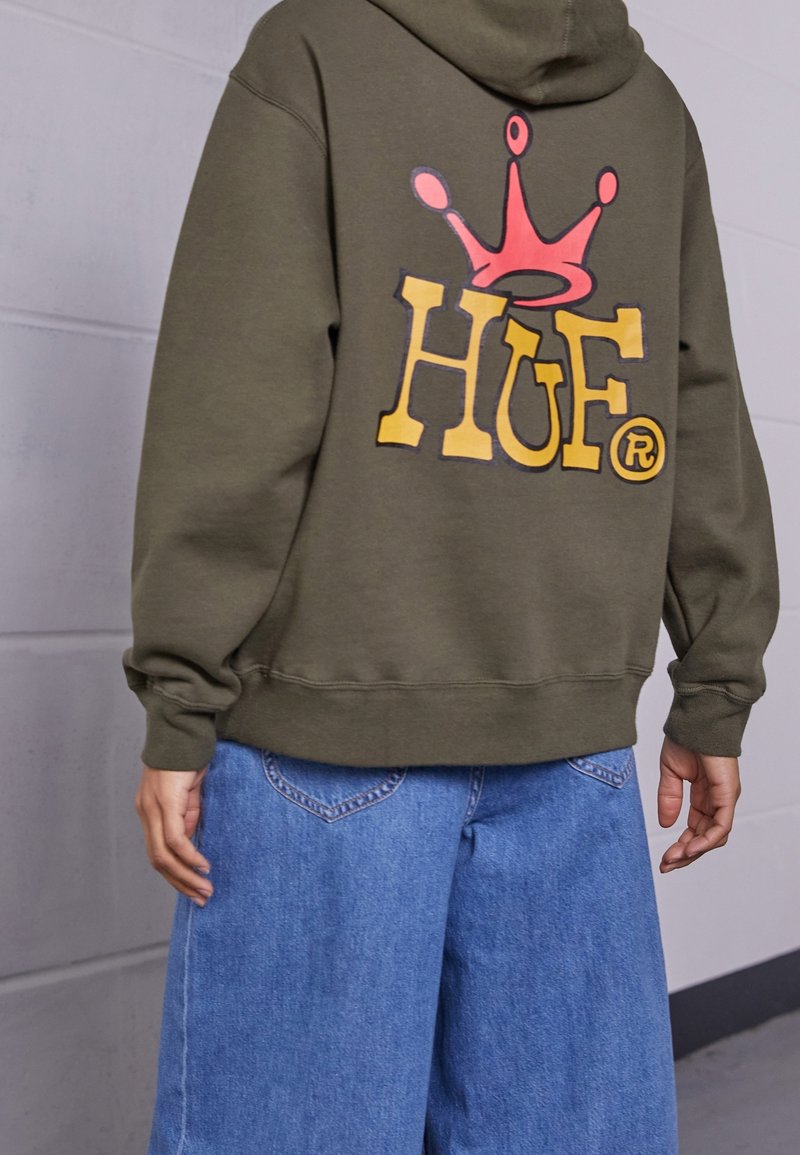 Felpa verde oliva con un grande logo stampato sul retro in giallo e rosso. Il materiale sembra morbido; maniche e orlo sono elasticizzati.