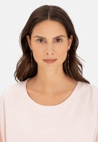 Goldfarbene Ohrringe mit schlanken, vertikalen Stäben und kleinen runden Akzenten. Das Modell trägt einen hellpinkfarbenen Sweatshirt mit runden Ausschnitt.