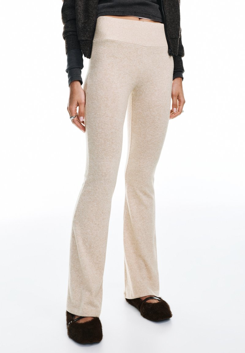 Pantalon évasé beige en tissu doux et texturé ; taille haute ; coupe mi-haute ; accompagné de chaussons marron pelucheux.