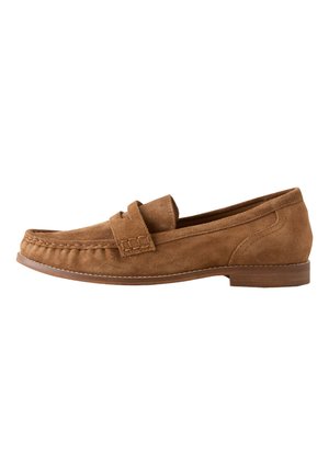Mocassins en daim marron portés avec un jean bleu à ourlet effiloché, debout sur un sol marron réfléchissant.