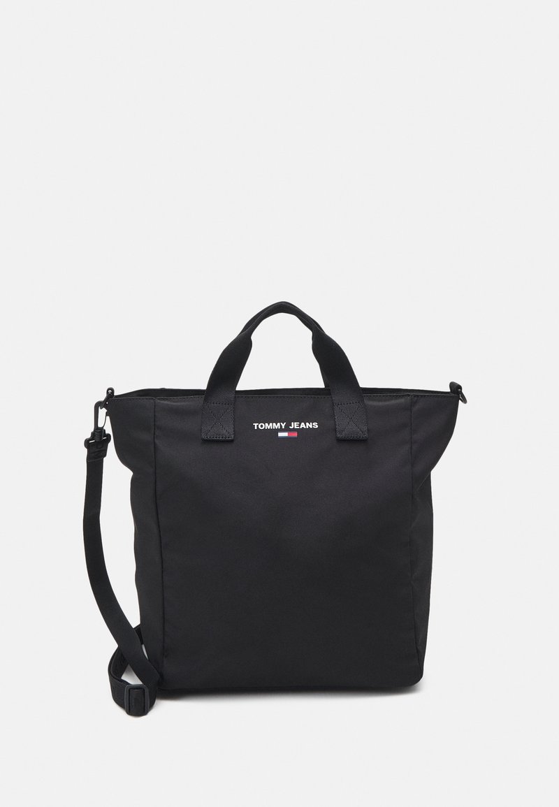 Sac cabas noir Tommy Jeans avec poignées supérieures et bandoulière réglable sur un fond clair uni.