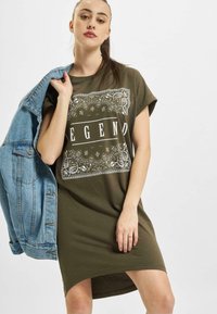 Oliwkowa sukienka w t-shircie z krótkimi rękawami, z białym napisem "LEGEND" oraz skomplikowanym wzorem graficznym, w połączeniu z jasną dżinsową kurtką.