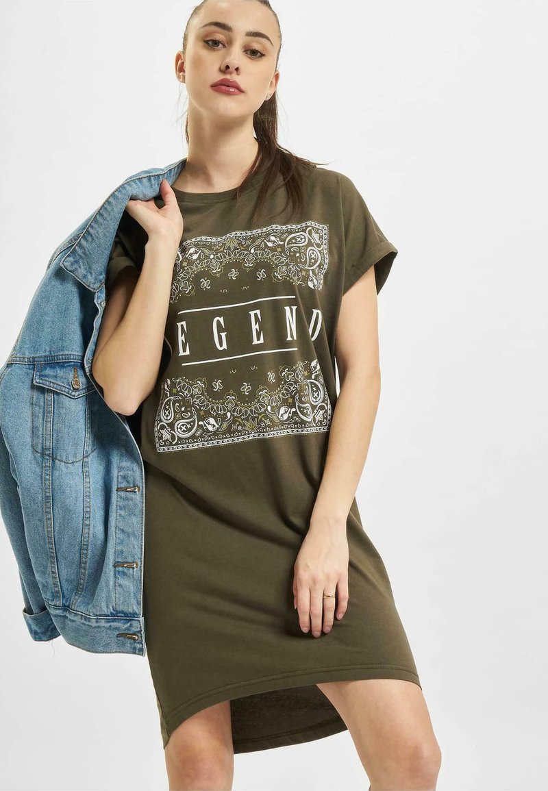 Oliwkowa sukienka w t-shircie z krótkimi rękawami, z białym napisem "LEGEND" oraz skomplikowanym wzorem graficznym, w połączeniu z jasną dżinsową kurtką.
