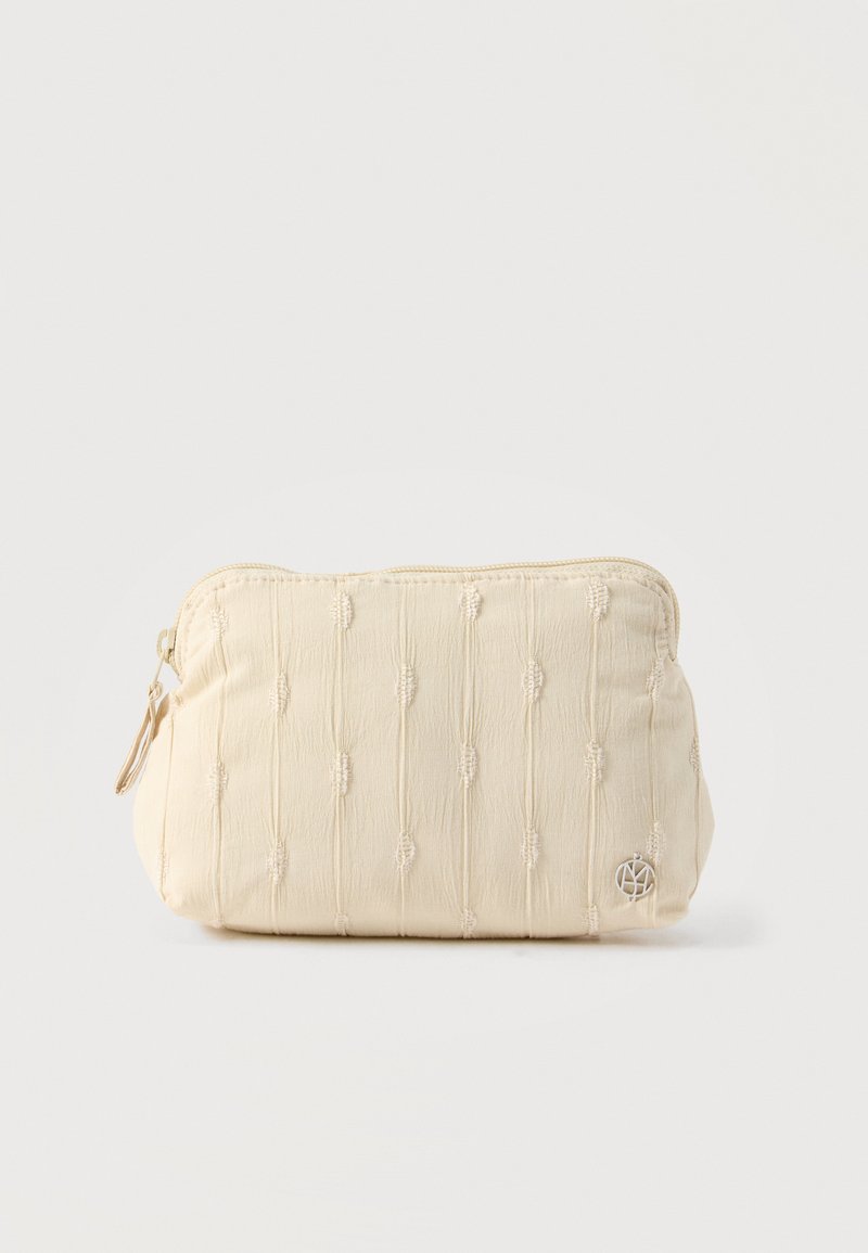 Κρεμ θήκη από ύφασμα με κάθετη υφασμάτινη ραφή, κλείσιμο με φερμουάρ και μικρό στρογγυλό μεταλλικό λογότυπο στην κάτω δεξιά γωνία.