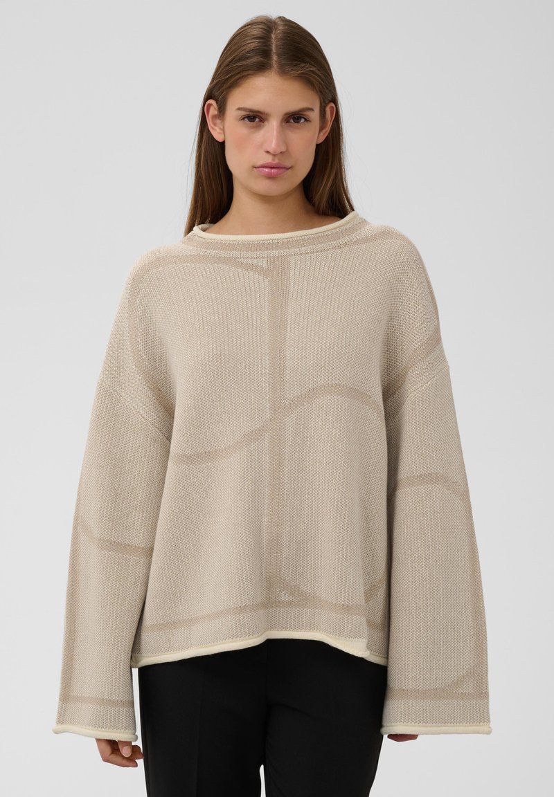 Jeune femme aux longs cheveux bruns portant un pull en maille beige ample et un pantalon noir, debout devant un fond clair uni.