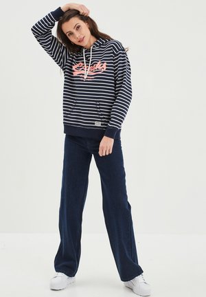 Felpa a righe navy e bianche con logo rosa, dotata di cappuccio con coulisse e tasca frontale, abbinata a pantaloni scuri a gamba larga e sneakers bianche.