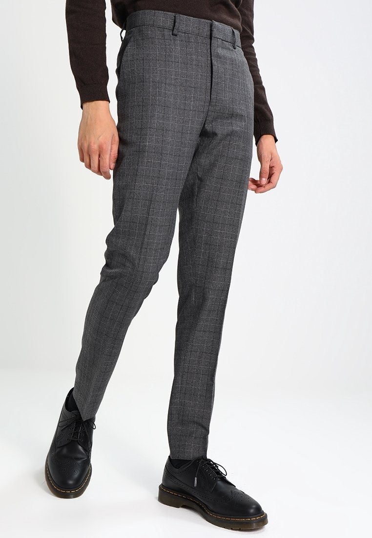 Personne portant un pantalon slim à carreaux gris et des chaussures en cuir noires à lacets, debout sur un fond blanc.