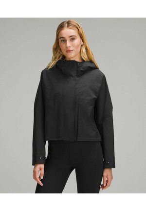 Frau mit langen blonden Haaren, die eine schwarze Kapuzenjacke und schwarze Leggings trägt, steht vor einem schlichten hellgrauen Hintergrund.