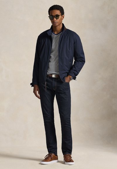 Ναυτικό ελαφρύ μπουφάν με γιακά που στέκεται όρθιος, ριγέ T-shirt από κάτω, σκούρα τζιν και καφέ αθλητικά παπούτσια με κορδόνια. Στρογγυλά γυαλιά ηλίου ολοκληρώνουν την εμφάνιση.