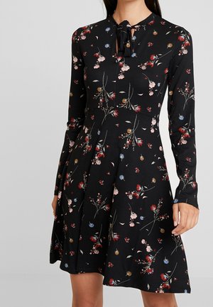 Zwarte jurk met lange mouwen met een multicolor bloemenprint en een strikdetail aan de voorkant bij de halslijn, gedragen door een staand persoon.
