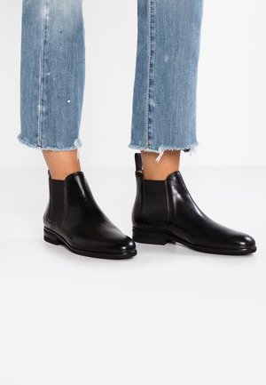 Bottes noires en cuir portées avec un jean bleu à ourlet effiloché, montrées de mi-mollet vers le bas sur un fond de sol blanc.