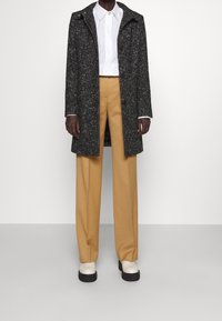 Mörk, prickig kappa, knäppt framtill, bärs över en vit skjorta och beige vida byxor. Enkla ljusa skor kompletterar outfiten.