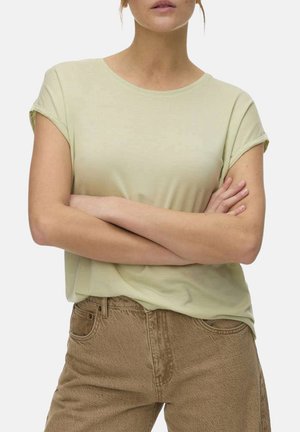 Femme les bras croisés portant une chemise à manches courtes vert clair rentrée dans un pantalon marron taille haute sur un fond uni.