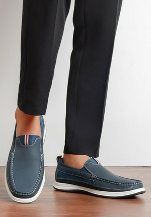 Chaussures décontractées à enfiler bleu marine avec semelles blanches et languettes rayées rouge, blanc et bleu, portées avec un pantalon noir sur un sol en bois.