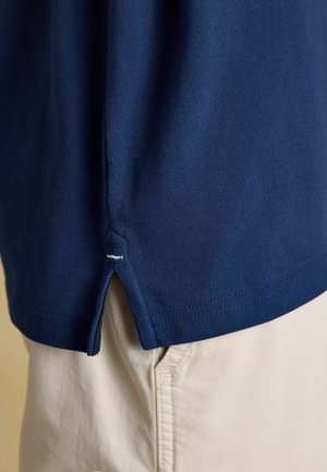 Detale rękawa granatowej koszuli z teksturowanej tkaniny na beżowych spodniach, pokazujące boczne rozcięcie z białym przeszyciem.