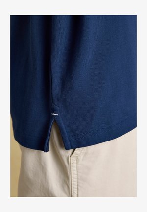 Detale rękawa granatowej koszuli z teksturowanej tkaniny na beżowych spodniach, pokazujące boczne rozcięcie z białym przeszyciem.