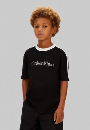 Fiatal fiú zsebében tartott kézzel, fekete, fehér részletekkel díszített Calvin Klein pólót viselve, egyszerű világosszürke háttér előtt áll.