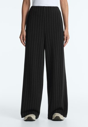 Pantalon large noir à rayures fines porté avec des baskets blanches et un haut blanc, vue de face de la taille aux pieds.