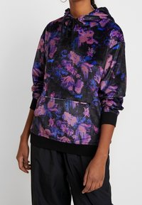 Kvinna iklädd en svart huvtröja med lila och blå blommönster, svarta dragkedjor, framficka och svarta byxor mot en enhetlig bakgrund.