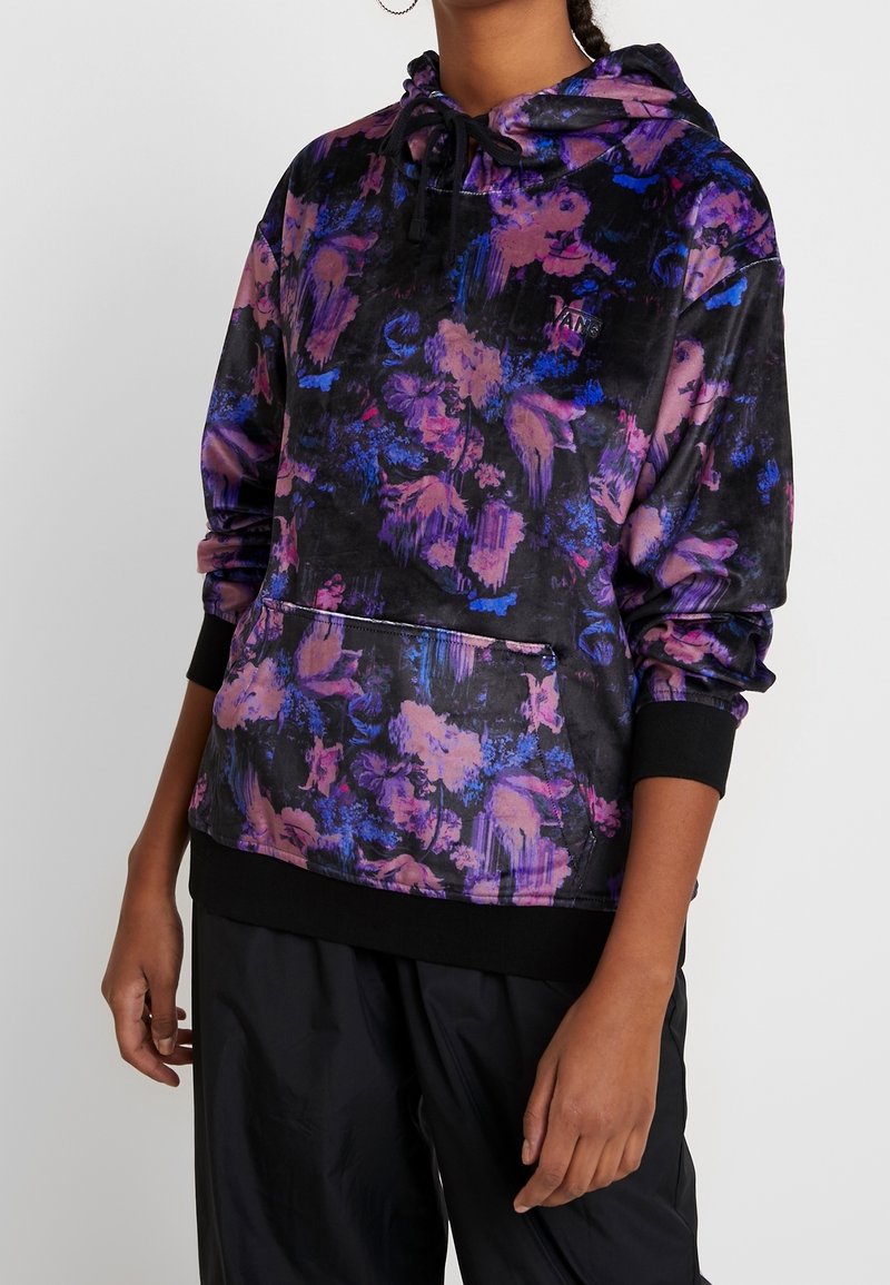 Kvinna iklädd en svart huvtröja med lila och blå blommönster, svarta dragkedjor, framficka och svarta byxor mot en enhetlig bakgrund.