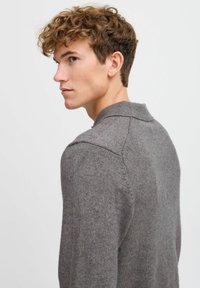 Grauer Strickpullover mit weicher Textur, der einen gerippten Kragen und Schulternaht hat. Modell seitlich gezeigt mit kurzen, lockigen Haaren.