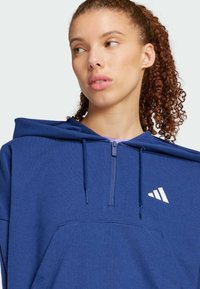 Plavi zip-up dukserica od mekanog materijala, s kenguruh džepom i bijelim logotipom na prsima. Kapuljača ima pertle za podešavanje.