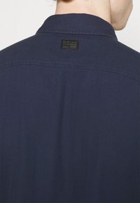 Maglietta blu navy con colletto a punta e un'etichetta nera sul retro. Presenta una texture liscia e cuciture pulite.