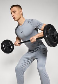 Camisa atlética gris y pantalones de tela suave, con un logo de Nike negro. El sujeto levanta una barra con pesas negras.