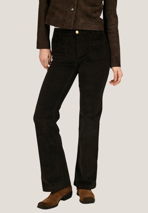 Femme portant une veste en daim marron foncé, un pantalon noir taille haute avec des poches avant et des bottines en daim marron.